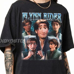 May include: Dark gray t-shirt featuring a graphic of Flynn Rider from the animated film Tangled. The design includes multiple images of the character with the name "Flynn Rider" in a blue, stylized font. The shirt has a vintage, distressed look.