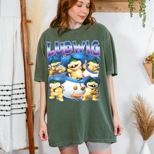Könnte beinhalten: Ein olivgrünes Kurzarm-T-Shirt mit einem Grafikdruck der Figur Ludwig aus der Super-Mario-Serie. Das Design zeigt den Namen LUDWIG in fetten lila Buchstaben über mehreren Illustrationen der Figur in verschiedenen Posen.