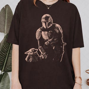May include: A dark brown short-sleeved t-shirt featuring a light-colored graphic print of a helmeted armored character standing next to a small, large-eared creature. The design is rendered in a textured, sketch-like style on the front of the shirt.