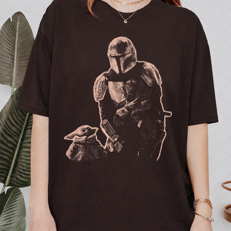 May include: A dark brown short-sleeved t-shirt featuring a light-colored graphic print of a helmeted armored character standing next to a small, large-eared creature. The design is rendered in a textured, sketch-like style on the front of the shirt.