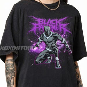 Könnte beinhalten: Schwarzes T-Shirt mit einer Grafik von Black Panther in einer hockenden Pose mit lila Energieeffekten. Die Worte "BLACK PANTHER" sind in einer stilisierten lila Schriftart dargestellt. Das Shirt hat einen Used-Look.