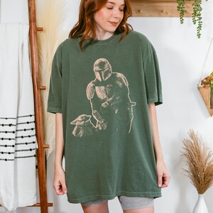May include: An olive green short-sleeve t-shirt featuring a light-colored graphic print of a helmeted warrior figure standing next to a small creature. The shirt has a crew neckline and a relaxed, loose-fitting silhouette with a straight hemline.