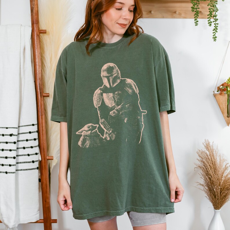 May include: An olive green short-sleeve t-shirt featuring a light-colored graphic print of a helmeted warrior figure standing next to a small creature. The shirt has a crew neckline and a relaxed, loose-fitting silhouette with a straight hemline.