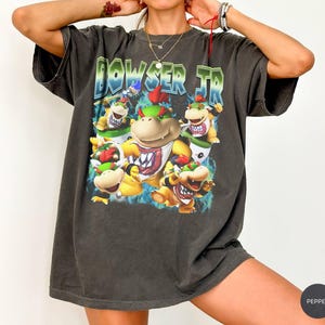Può includere: Una t-shirt a maniche corte grigio antracite con una stampa grafica del personaggio Bowser Jr. in varie pose. Il testo "BOWSER JR" appare in un carattere audace e stilizzato sopra le illustrazioni del personaggio, su uno sfondo di fiamme blu.