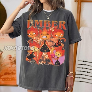 May include: Dark gray t-shirt featuring the word "EMBER" in orange lettering above a graphic of cartoon fire characters. The characters have orange flames for hair and are wearing black dresses. The shirt is a relaxed fit.