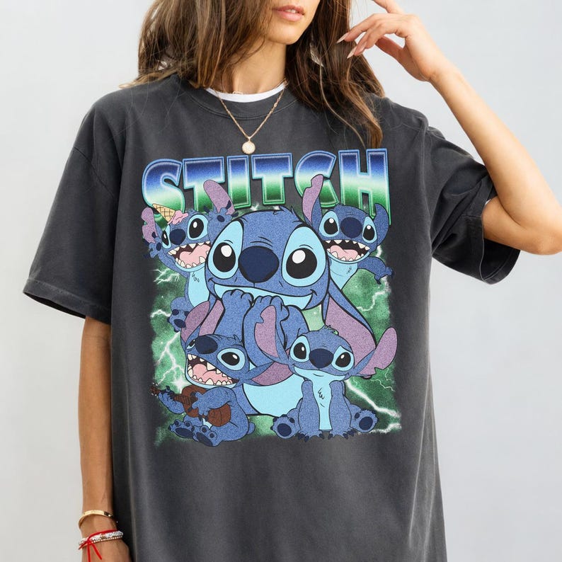 May include: A charcoal gray short-sleeve t-shirt featuring a graphic print of the character Stitch in various poses. The design includes the word STITCH in a blue and green gradient font, lightning bolts, and a small ice cream cone graphic.