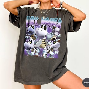 May include: A charcoal gray short-sleeve t-shirt featuring a graphic print of multiple white skeletal turtle characters. The text DRY BONES is printed in a metallic blue and purple gradient font above the characters, set against a purple background.