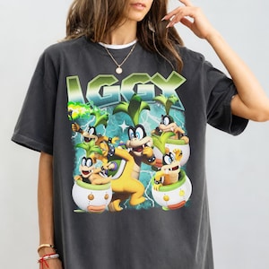 Può includere: Una t-shirt a maniche corte color grigio antracite con una stampa grafica del personaggio Iggy Koopa della serie Super Mario. Il design include il testo IGGY in grandi lettere verdi sopra diverse rappresentazioni del personaggio.