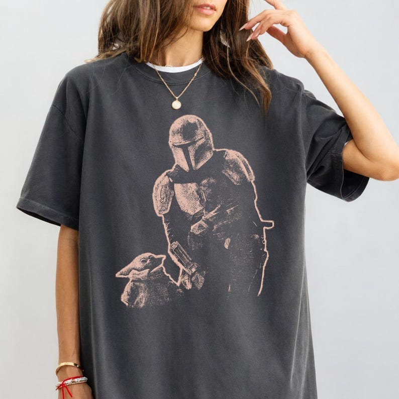 May include: A charcoal gray short-sleeve t-shirt featuring a light pink graphic print of a helmeted armored character standing next to a small creature with large ears. The shirt has a crew neckline and a relaxed fit, showcasing a detailed illustration.