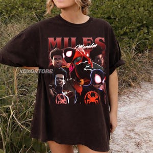 K&ouml;nnte beinhalten: Dunkelbraunes T-Shirt mit einer Grafik von Miles Morales und anderen Charakteren aus dem Spider-Man-Universum. Das Shirt tr&auml;gt den Text "MILES Morales" im Vintage-Stil. Der Markenname "XOXOSTORE" ist ebenfalls sichtbar.