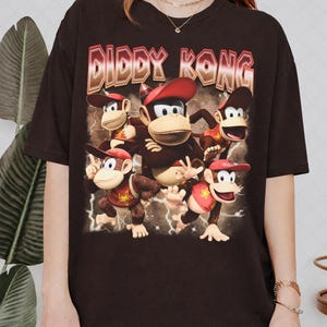 May include: A dark brown short-sleeved t-shirt featuring a large graphic print of Diddy Kong in various poses. The text DIDDY KONG appears in a bold, metallic-style font at the top, with lightning bolt accents surrounding the characters.