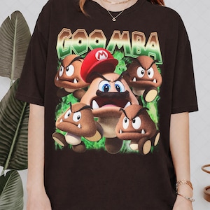 Peut inclure: Un t-shirt noir à manches courtes avec une impression graphique du mot GOOMBA en lettres vertes et brunes au-dessus d'un personnage central portant un chapeau rouge, entouré de quatre créatures brunes en forme de champignon sur fond de fumée verte.