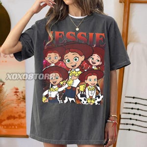Op de afbeelding: Donkergrijs T-shirt met een afbeelding van het personage Jessie uit Toy Story. Het woord "JESSIE" staat in oranje boven de afbeelding. De afbeelding toont Jessie in verschillende poses.