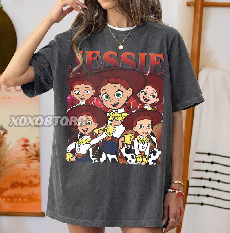 Op de afbeelding: Donkergrijs T-shirt met een afbeelding van het personage Jessie uit Toy Story. Het woord "JESSIE" staat in oranje boven de afbeelding. De afbeelding toont Jessie in verschillende poses.