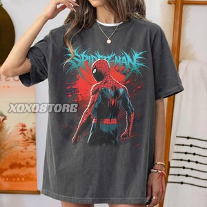 May include: Dark gray t-shirt featuring a graphic of Spider-Man from the back, with red and blue suit details. The text "SPIDER-MAN" is in blue, with a red background. The shirt has the text "XOXOSTORE" on the left side.