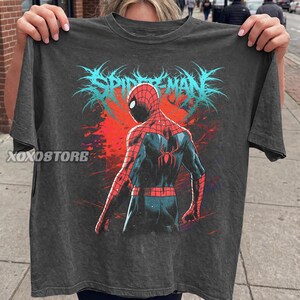 May include: Dark gray t-shirt featuring a graphic of Spider-Man from the back, with a red and blue suit, set against a red and blue background. The word "SPIDER-MAN" is printed in blue above the character. The brand name "XOXOSTORE" is printed on the left side of the shirt.