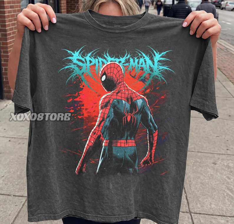 May include: Dark gray t-shirt featuring a graphic of Spider-Man from the back, with a red and blue suit, set against a red and blue background. The word "SPIDER-MAN" is printed in blue above the character. The brand name "XOXOSTORE" is printed on the left side of the shirt.