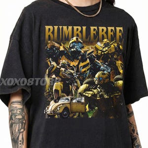 May include: Black t-shirt featuring a graphic of Bumblebee from Transformers. The design includes the word "BUMBLEBEE" in gold lettering, along with images of the character and a yellow car. The shirt is a dark gray color.