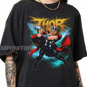 May include: Dark gray t-shirt featuring a graphic of Thor, wielding his hammer. The design includes the word "Thor" in gold, with a red cape and blue background. The shirt has a vintage, worn look.