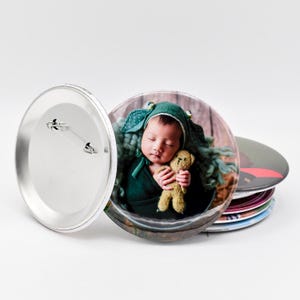 May include: A stack of circular pin-back buttons with a metallic silver backing and a pin mechanism. The top button features a high-resolution color photograph of a sleeping infant wearing a green bonnet and holding a small tan teddy bear.