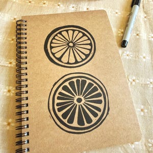 Peut inclure: Un cahier à spirales avec une couverture en papier kraft ornée de deux motifs de tranches d'agrumes noirs. Un marqueur noir repose sur la surface à motifs floraux à côté du cahier. Le cahier est ouvert, affichant le motif de la couverture.