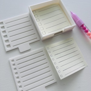 May include: A white notepad set with a matching pen. The set includes a small rectangular box holding lined paper, two white dividers, and a pink pen with decorative beads. The notepad paper has check boxes and lines.