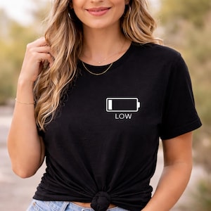 Pode incluir: T-shirt preta com gola redonda, com um ícone de bateria branco e a palavra "LOW" impressa abaixo. A camisa é feita de material macio, adequada para uso casual.