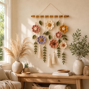 May include: A wall hanging featuring crocheted flowers in shades of pink, yellow, purple, and cream, suspended from a horizontal wooden branch by white cords. The arrangement includes green crocheted leaves, small wooden beads, and white tassels.