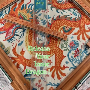 May include: A Mahjong set featuring a clear plastic storage tube with a green bird logo and the text MAHJ MUST HAVES. The game includes wooden racks and tiles with a mottled brown pattern, arranged on a mat with a large orange dragon illustration.