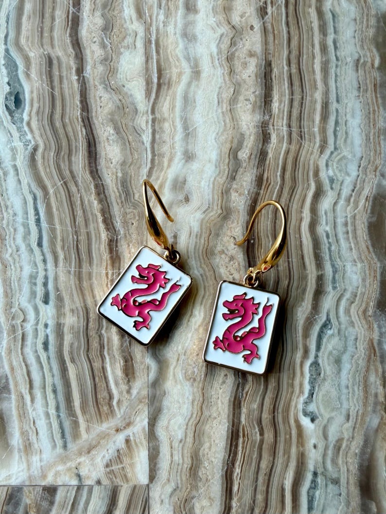 Op de afbeelding: Een paar goudkleurige oorbellen met een rechthoekige witte emaille hanger. Elke hanger heeft een roze drakenontwerp en een goudkleurige haak. De oorbellen worden gepresenteerd op een gestructureerd, gemarmerd oppervlak.