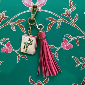 May include: A gold-toned keychain with a pink tassel and a small, rectangular charm. The charm features a bird on a branch design. The background is a teal fabric with a floral pattern.