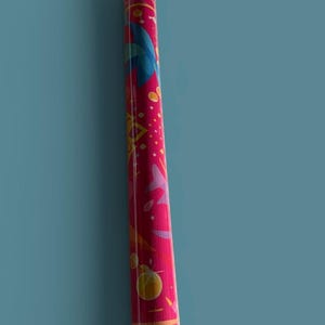 May include: A cylindrical storage tube with a vibrant magenta background featuring a colorful graphic pattern of blue, yellow, and pink shapes. The top and bottom caps are white, and a small green label with the text MAHI is visible near the top.