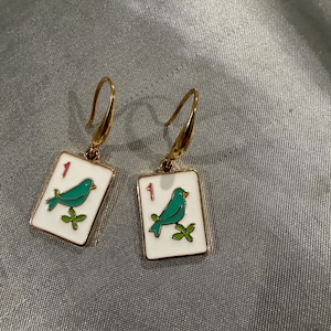 May include: A pair of gold-colored earrings. Each earring features a white rectangular charm with a gold border. The charm has a teal bird perched on a green branch, with the number "1" in red. The earrings have a hook closure.