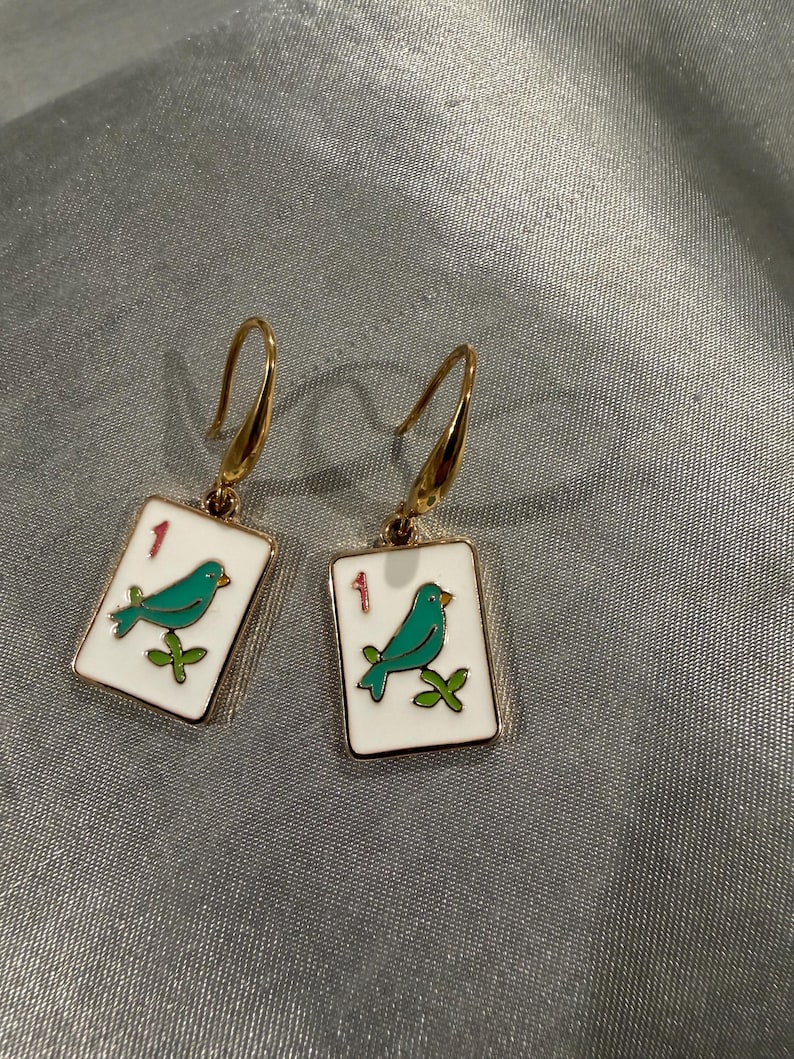 May include: A pair of gold-colored earrings. Each earring features a white rectangular charm with a gold border. The charm has a teal bird perched on a green branch, with the number "1" in red. The earrings have a hook closure.