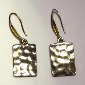 May include: A pair of gold-toned earrings. Each earring features a rectangular, textured pendant suspended from a curved ear wire. The pendants have a hammered metal appearance, reflecting light. The earrings are designed for pierced ears.