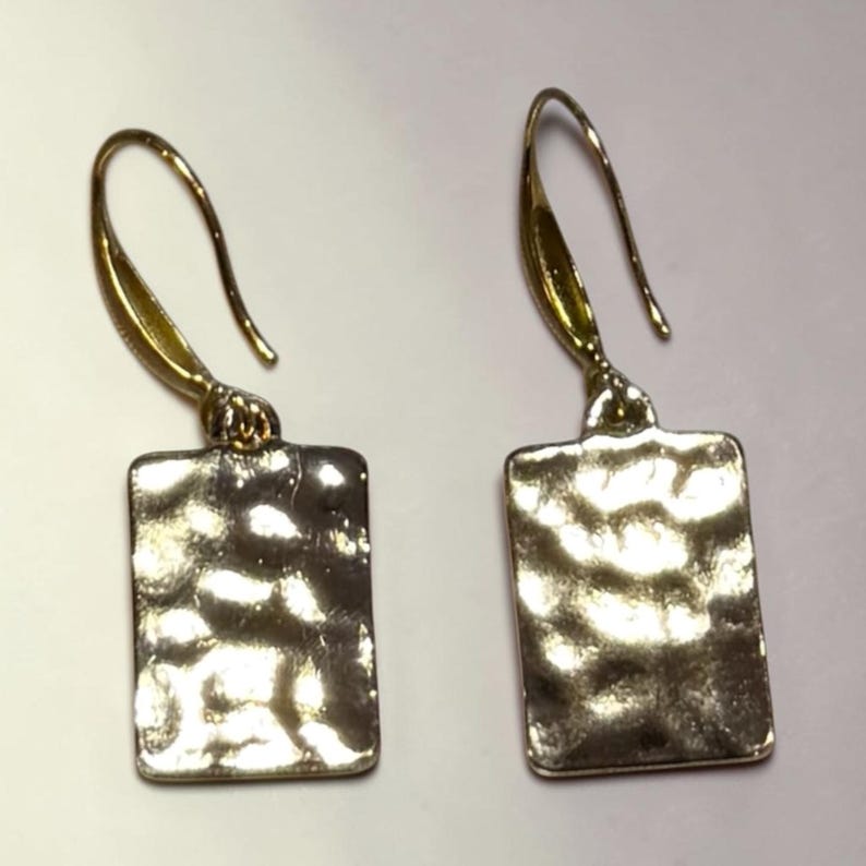May include: A pair of gold-toned earrings. Each earring features a rectangular, textured pendant suspended from a curved ear wire. The pendants have a hammered metal appearance, reflecting light. The earrings are designed for pierced ears.