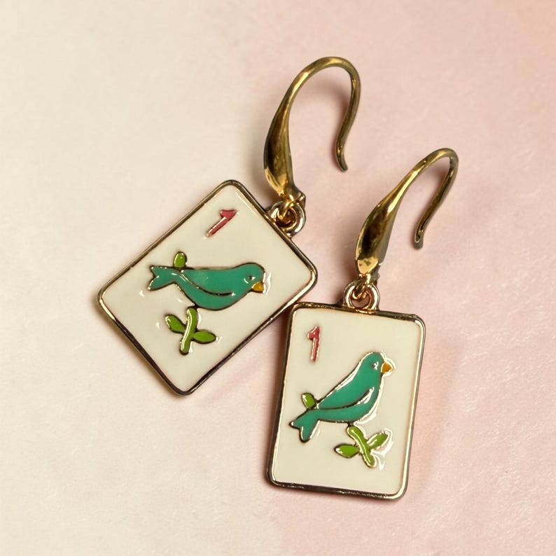 May include: A pair of gold-tone earrings. Each earring features a white rectangular charm with a green bird illustration, a red number 1, and a gold border. The bird is perched on a green branch. The earrings have a hook closure.
