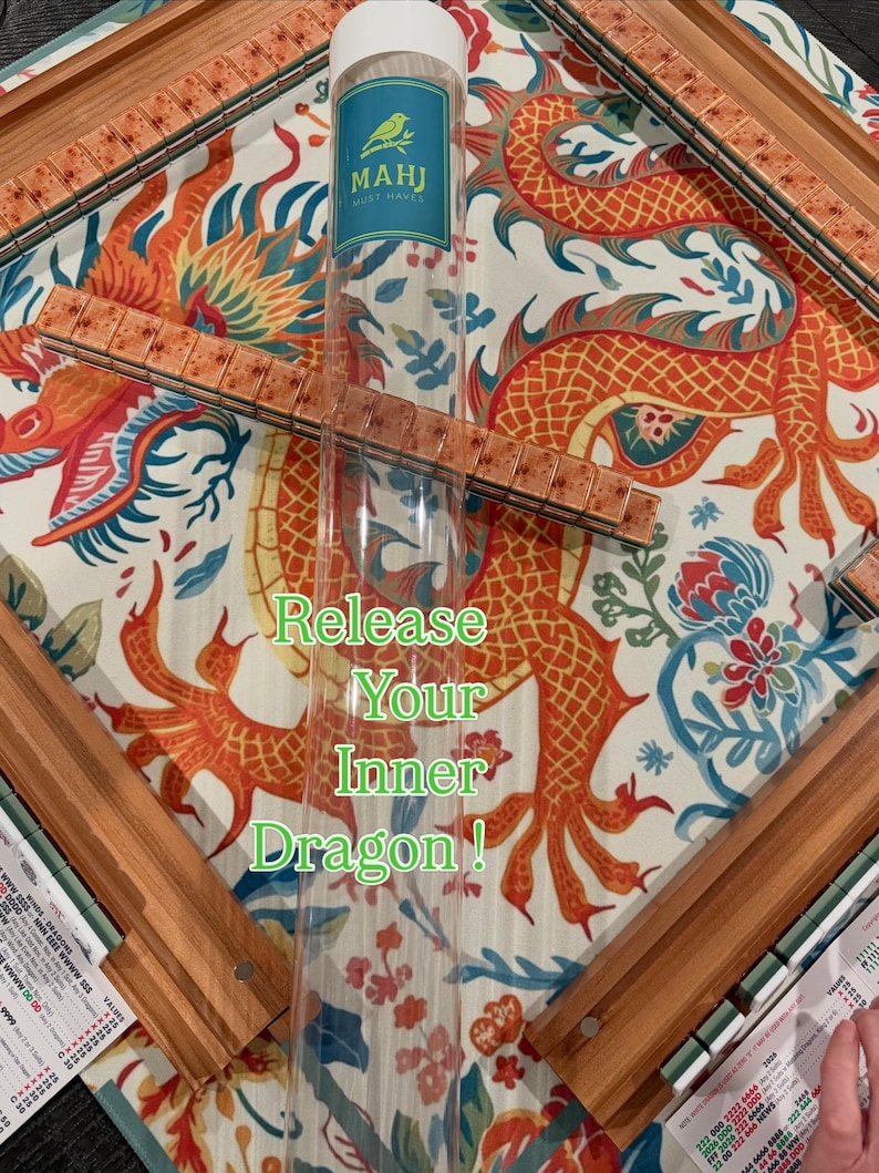 May include: A Mahjong set featuring a clear plastic storage tube with a green bird logo and the text MAHJ MUST HAVES. The game includes wooden racks and tiles with a mottled brown pattern, arranged on a mat with a large orange dragon illustration.