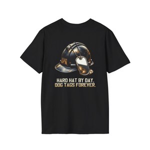 Puede incluir: Camiseta negra con un gráfico de un casco y placas de identificación. El diseño incluye el texto "HARD HAT BY DAY, DOG TAGS FOREVER." La imagen tiene un aspecto vintage y desgastado.