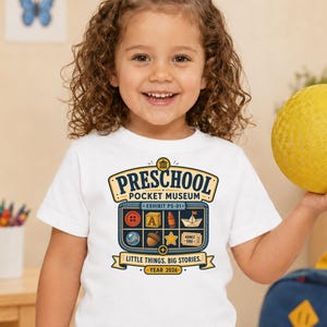 May include: A white short-sleeved t-shirt featuring a graphic print that reads Preschool Pocket Museum, Exhibit PS-01, Little Things. Big Stories. Year 2026. The design includes icons of a button, block, crayon, boat, marble, acorn, star, and ticket.