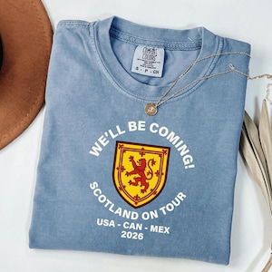 A blue t-shirt with a graphic design and text, along with a brown hat and some dried leaves, are displayed on a white surface.