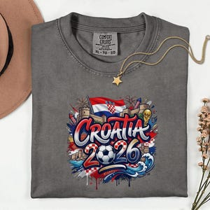 a gray t-shirt with a graphic design depicting the word "Croatia" surrounded by various elements associated with the country, such as a flag, buildings, and a soccer ball. The design also includes the text "Croatia 2026" and a star. The t-shirt is placed on a white surface, with a hat and some flowers visible in the background.