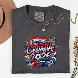 a gray t-shirt with a graphic design depicting the word "Croatia" surrounded by various elements associated with the country, such as a soccer ball, a flag, and a castle. The design also includes the text "Croatia 2026" and a star. The t-shirt is placed on a white surface, with a hat and some flowers visible in the background.