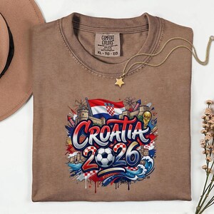 a brown t-shirt with a graphic design depicting the word "Croatia" surrounded by various elements such as a soccer ball, flags, and buildings.