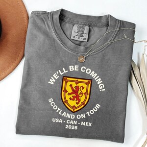 a gray t-shirt with a red and yellow shield design, along with the text "WE'LL BE COMING! SCOTLAND ON TOUR USA - CAN - MEX 2026".