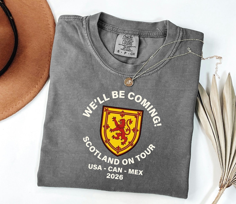 a gray t-shirt with a red and yellow shield design, along with the text "WE'LL BE COMING! SCOTLAND ON TOUR USA - CAN - MEX 2026".