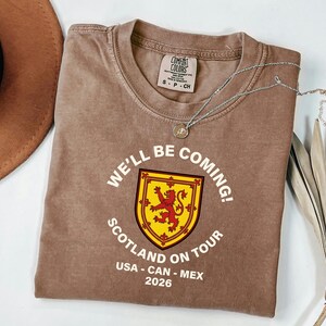 a brown t-shirt with a graphic design, a necklace, a hat, and some leaves on a white background.