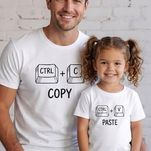 May include: Two white t-shirts with black graphics. One says "CTRL + C COPY", the other "CTRL + V PASTE". A man and a child are wearing the shirts, smiling. The background is a white brick wall.