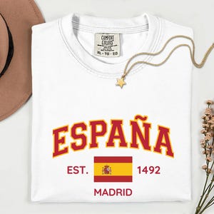 May include: A white short-sleeved t-shirt featuring the red and yellow text ESPAÑA in an arched font. Below the text is the Spanish flag, the text EST. 1492, and the word MADRID in red. The shirt has a Comfort Colors label indicating size XL.