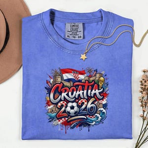 a blue t-shirt with a graphic design depicting the word "Croatia" surrounded by various elements such as a soccer ball, flags, and buildings. The t-shirt is laid out on a white surface, with a hat and some flowers nearby.